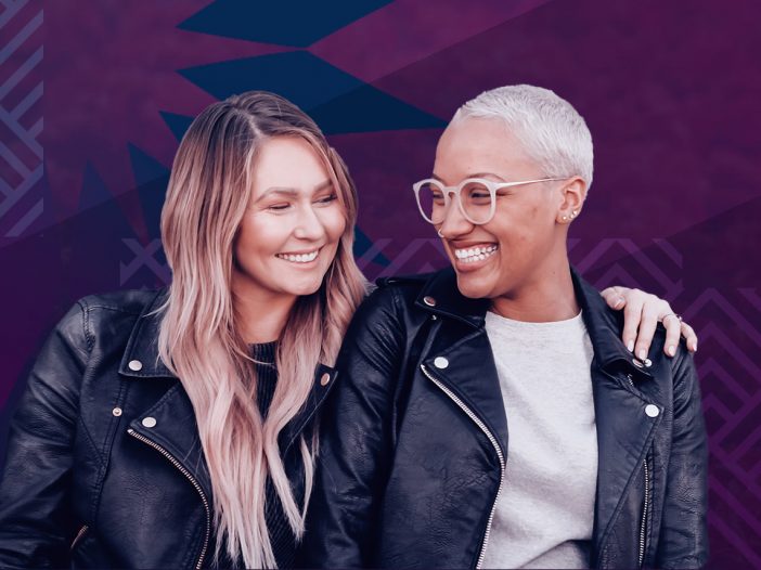 Caucasian female with long blond hair puts her arm around an African American female with short silver hair, they are smiling and looking at one another